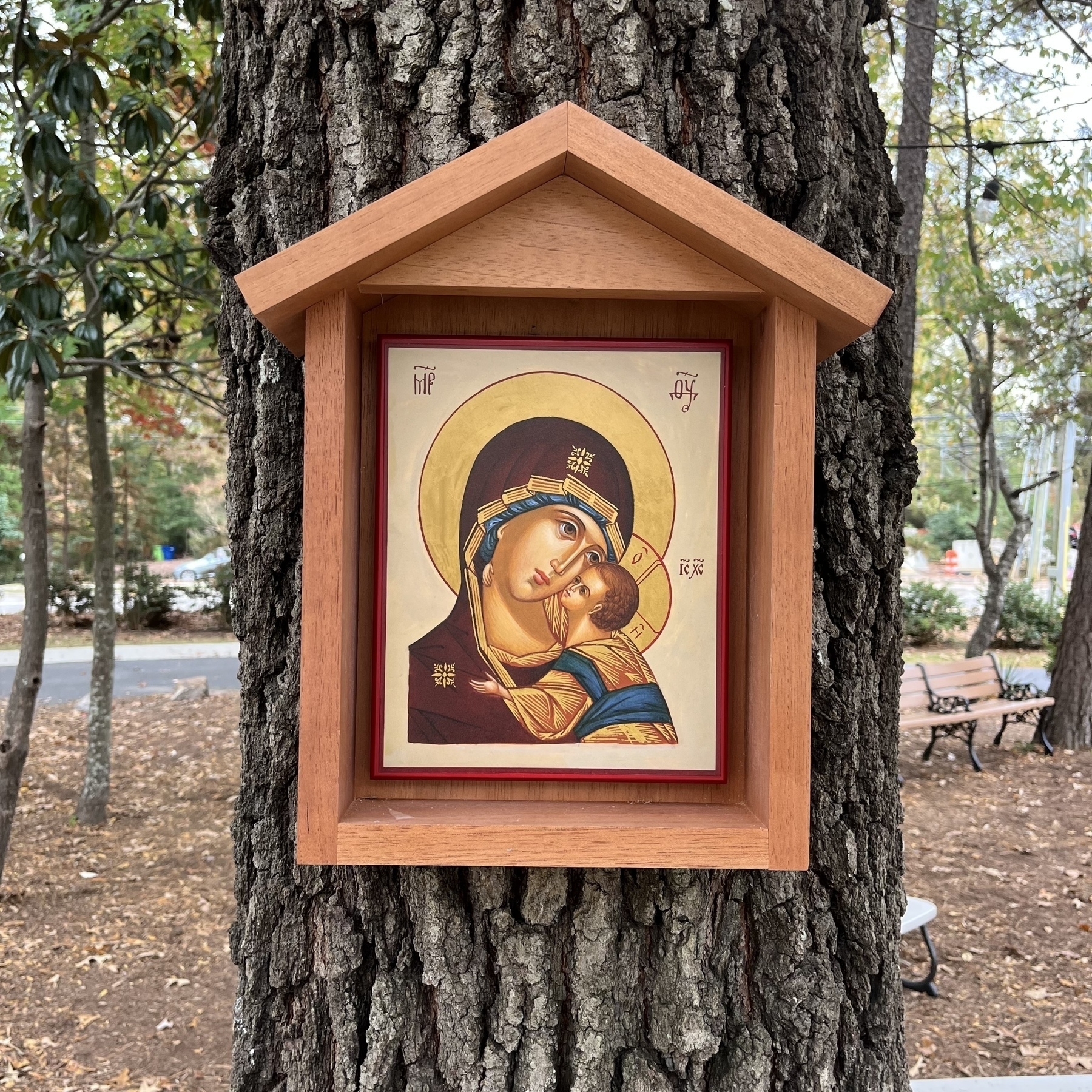 A religious icon of the Virgin Mary and Child is mounted on a tree within a wooden frame, surrounded by a serene outdoor setting.